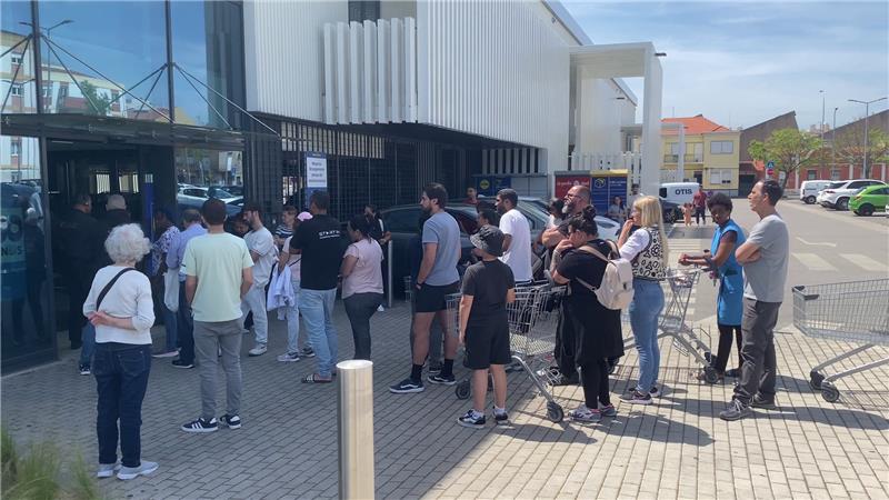 Mensen wachten in de zon met winkelwagens voor de ingang van een supermarkt of winkelpand.'